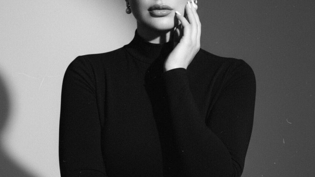 Woman dressed in a black turtleneck, touching her face, photographed in a minimal studio setting.